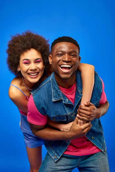 Cheerful young african american woman with bold makeup in sundress embracing stylish best friend in denim vest and looking at camera isolated on blue, fashionable friends in trendy clothes — Stock Photo