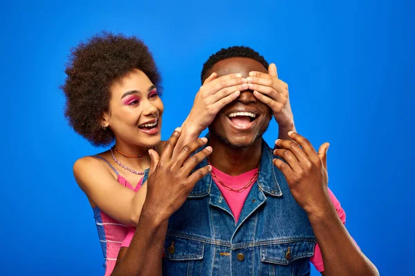 Positiva joven afroamericana mujer con audaz maquillaje que cubre los ojos al mejor amigo con estilo en chaleco de mezclilla y de pie juntos aislados en azul, amigos de moda en ropa de moda - foto de stock
