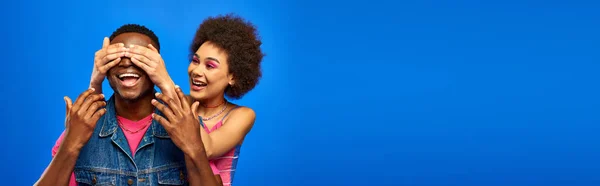 Giovane donna afroamericana sorridente con trucco luminoso che copre gli occhi del migliore amico alla moda in gilet di denim e in piedi isolato su amici blu e alla moda in abiti alla moda, banner — Foto stock