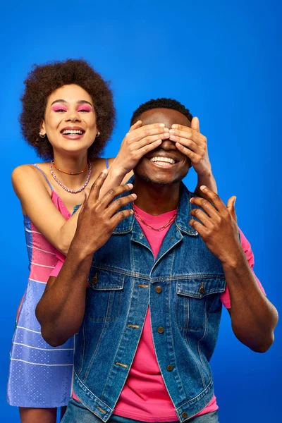 Positive junge afrikanisch-amerikanische Frau mit kühnem Make-up, die die Augen ihrer besten Freundin im Sommeroutfit bedeckt, während sie isoliert auf blauen, modischen Freundinnen in trendigen Klamotten steht — Stockfoto