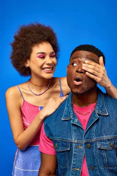 Jovem mulher americana africana alegre com maquiagem ousada e sundress cobrindo o olho para o melhor amigo chocado na roupa de verão, enquanto estava isolado em azul, amigos da moda em roupas da moda — Fotografia de Stock