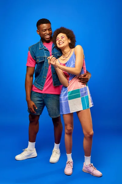 Full length of cheerful and stylish young african american man in denim vest and bright t-shirt hugging best friend in sundress on blue background, fashionable friends in trendy clothes — Stock Photo