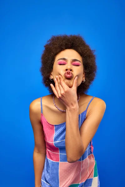 Portrait of stylish young african american woman with bold makeup and sundress closing eyes and pouting lips while posing and standing isolated on blue, charismatic model in summer outfit — Stock Photo