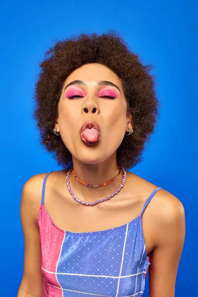 Portrait of young and stylish african american woman with bold makeup wearing summer sundress and sticking out tongue while standing isolated on blue, charismatic model in summer outfit — Stock Photo