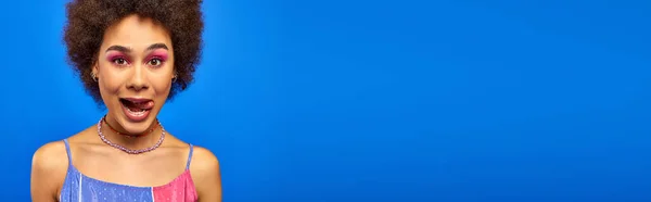 Giovane e trendy donna afroamericana con trucco audace e capelli naturali sporgenti lingua e guardando la fotocamera isolata su blu, modello carismatico in abito estivo, banner — Foto stock