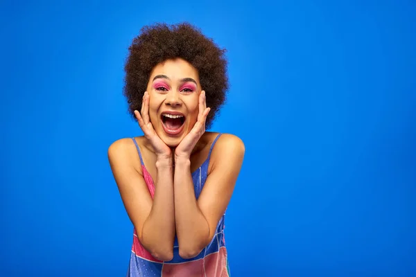 Excited young african american woman with bold makeup wearing sundress while touching cheeks and looking at camera and standing isolated on blue, charismatic model in summer outfit — Stock Photo