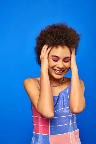 Portrait de jeune femme afro-américaine élégante et souriante aux cheveux naturels et au maquillage audacieux touchant la tête tout en se tenant debout et posant isolée sur un modèle bleu charismatique en tenue d'été — Photo de stock