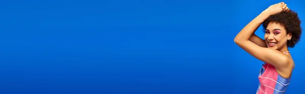 Allegro giovane donna afro-americana con trucco luminoso indossa sundress estate e guardando la fotocamera mentre in piedi isolato su blu, modello carismatico in abito estivo, banner — Foto stock