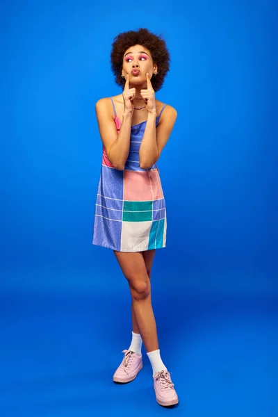 Full length of stylish young african american woman with bold makeup wearing sneakers and sundress and pouting lips while standing on blue background, charismatic model in summer outfit — Stock Photo
