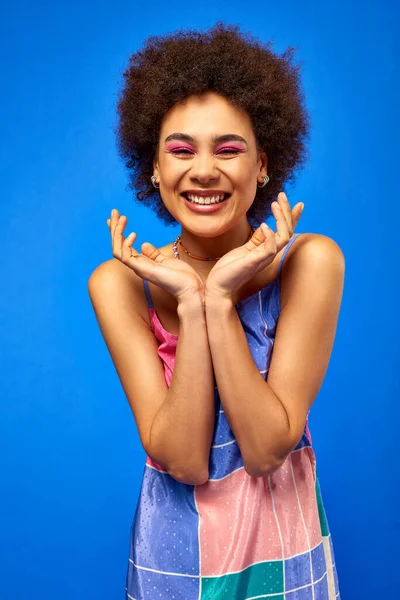 Retrato de mujer afroamericana joven y positiva con cabello natural y maquillaje audaz usando vestido de verano y posando mientras está de pie aislado en azul, modelo carismático en traje de verano - foto de stock