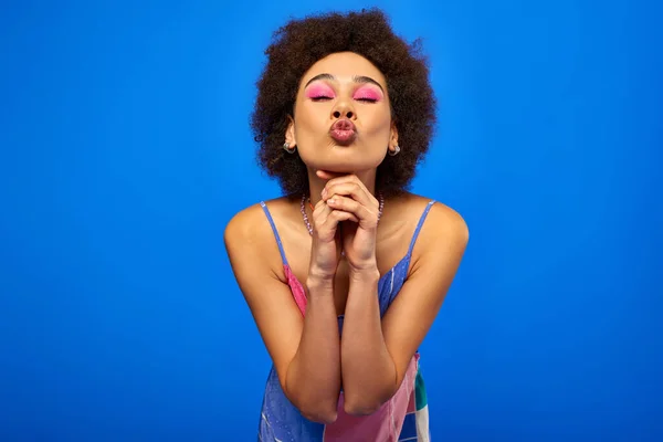 Retrato de una joven afroamericana con un elegante maquillaje audaz que lleva vestido de sol y labios de puchero mientras posa y está de pie aislado en azul, modelo carismático en traje de verano - foto de stock