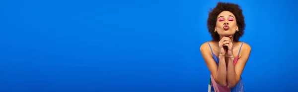 Молодая африканская американка с смелым макияжем и натуральными волосами, позирующая в сарафане и дующиеся губы, стоящая изолированная на синем с копировальным пространством, харизматичная модель в летнем наряде, баннер — стоковое фото