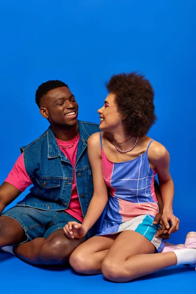Jóvenes y elegantes mejores amigos afroamericanos en trajes de verano sonriendo mientras se miran, pasando tiempo juntos y sentados en un fondo azul, amigos elegantes posando con confianza - foto de stock