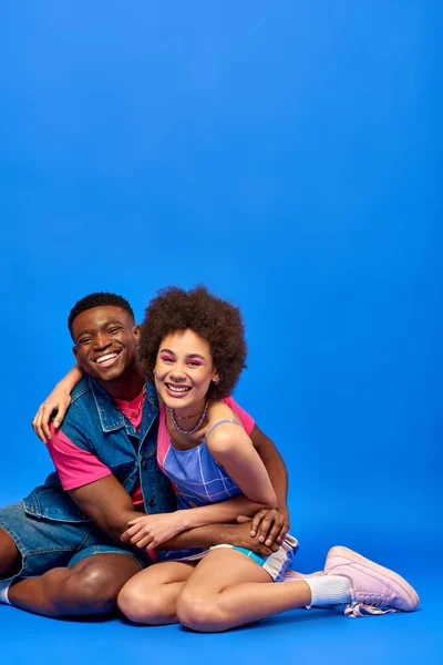 Улыбающийся молодой африканский американец в джинсовом жилете обнимает лучшего друга с смелым макияжем и стильным сарафаном и сидит вместе на синем фоне, стильные друзья уверенно позируют — стоковое фото