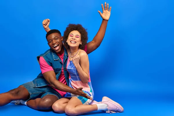Jovens amigos afro-americanos alegres em roupas de verão brilhantes acenando a mão e mostrando sim gesto na câmera enquanto sentados juntos no fundo azul, amigos elegantes posando com confiança — Fotografia de Stock