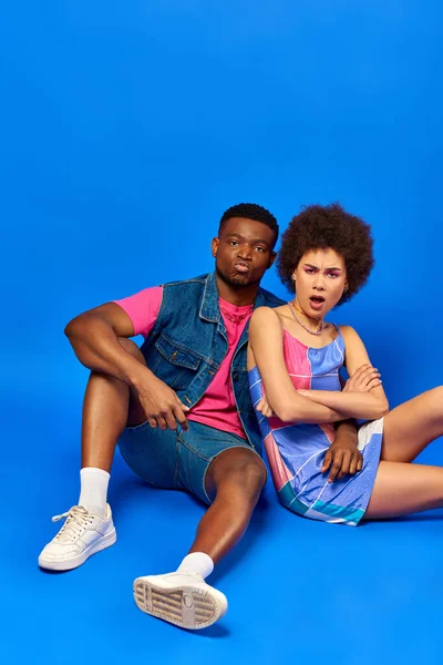 Offensé jeunes meilleurs amis afro-américains en tenues d'été lumineux regardant la caméra et croisant les bras tout en étant assis ensemble sur fond bleu, amis élégants posant en toute confiance — Photo de stock