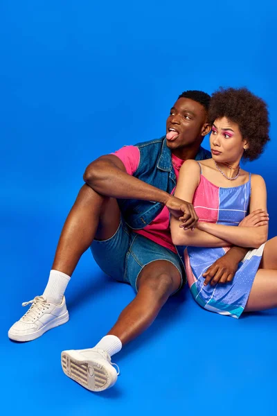 Offended young african american best friends in bright summer outfits crossing arms and sticking out tongue while sitting together on blue background, stylish friends posing confidently — Stock Photo