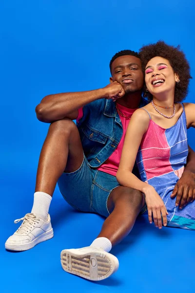 Молодий і стильний афроамериканський чоловік в літньому вбранні посміхається, сидячи біля веселого кращого друга в літній сукні, позуючи на синьому фоні, стильні друзі впевнено позують — стокове фото