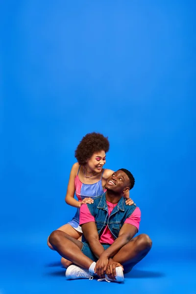 Smiling young african american woman with bright makeup wearing summer dress and hugging best friend in denim vest while sitting on blue background, stylish friends posing confidently — Stock Photo