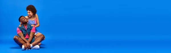 Sorridente giovane donna afroamericana con trucco audace abbracciando elegante migliore amico in t-shirt luminosa e gilet di jeans mentre seduto su sfondo blu, amici alla moda in posa con fiducia, banner — Foto stock