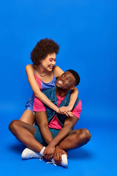 Mujer afroamericana joven positiva con cabello natural y maquillaje audaz abrazando mejor amigo en traje de verano de moda mientras está sentado en el fondo azul, amigos elegantes posando con confianza - foto de stock