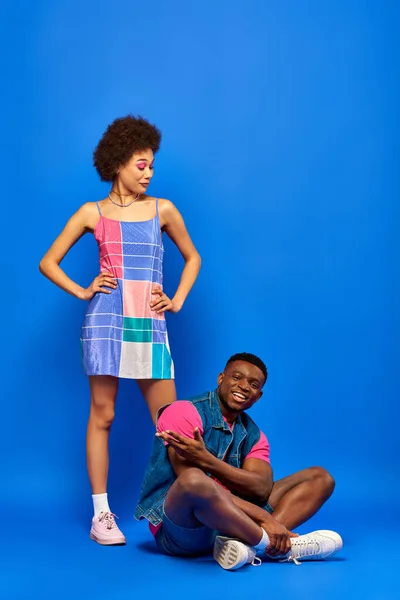 Full length of astonished young african american woman with bold makeup and sundress looking at stylish best friend smiling and sitting on blue background, stylish friends posing confidently — Stock Photo