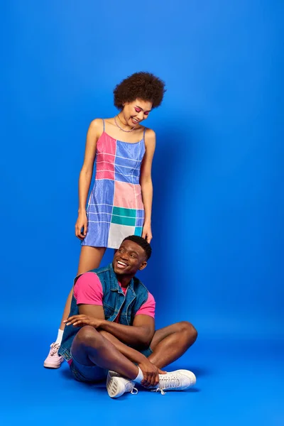 Pleine longueur de joyeux jeune homme afro-américain en tenue d'été assis près du meilleur ami en robe de soleil debout sur fond bleu, amis élégants posant en toute confiance, concept d'amitié — Photo de stock