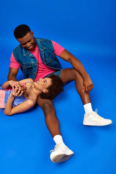 High angle view of positive young african american woman with natural hair and bold makeup lying near best friend in denim vest on blue background, stylish friends posing confidently — Stock Photo