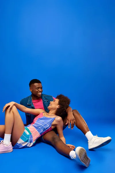 Positive young african american woman with bold makeup wearing sundress while posing with best friend in summer outfit and sitting on blue background, stylish friends posing confidently — Stock Photo