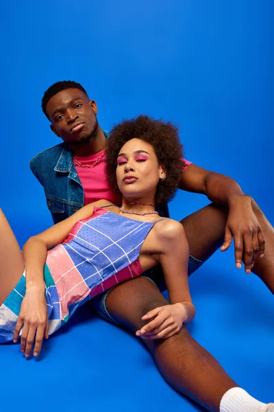 Moda e confiante jovens amigos afro-americanos em roupas de verão olhando para a câmera enquanto se sentam juntos no fundo azul, besties na moda irradiando confiança — Fotografia de Stock