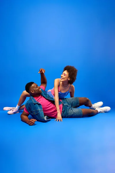 Positif jeune femme afro-américaine avec un maquillage audacieux en robe de soleil posant près élégant meilleur ami en gilet en denim et t-shirt sur fond bleu, besties à la mode rayonnant de confiance — Photo de stock