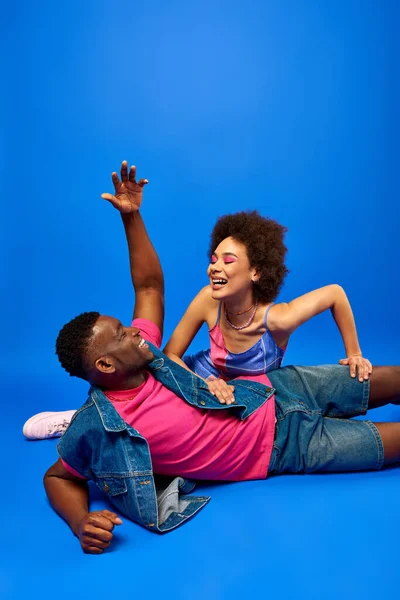 Cheerful young african american man with modern hairstyle talking to best friend with bold makeup and sundress while sitting on blue background, fashionable besties radiating confidence — Stock Photo