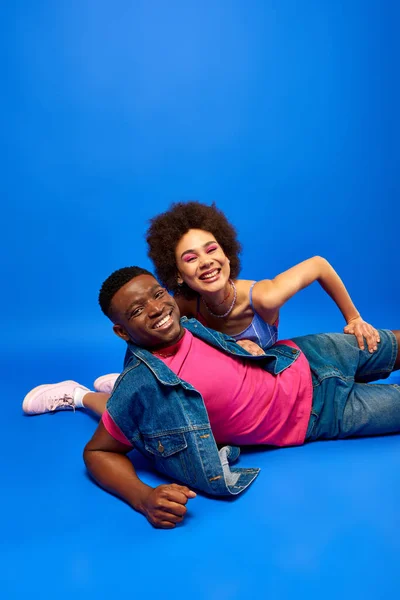 Joyeux jeune femme afro-américaine avec un maquillage audacieux regardant la caméra tout en posant près de meilleur ami à la mode en gilet en denim sur fond bleu, besties à la mode rayonnant de confiance — Photo de stock