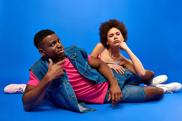 Pensif jeune afro-américain meilleurs amis dans des tenues d'été lumineuses et à la mode en regardant la caméra tout en étant couché ensemble sur fond bleu, besties à la mode rayonnant de confiance, amitié — Photo de stock