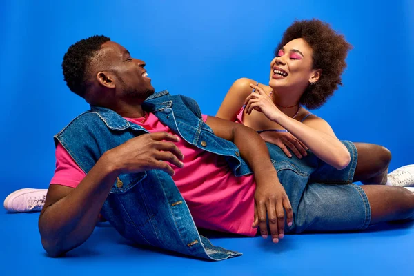 Souriant jeune femme afro-américaine avec un maquillage lumineux couché près du meilleur ami élégant en t-shirt et gilet en denim sur fond bleu, besties à la mode rayonnant de confiance — Photo de stock