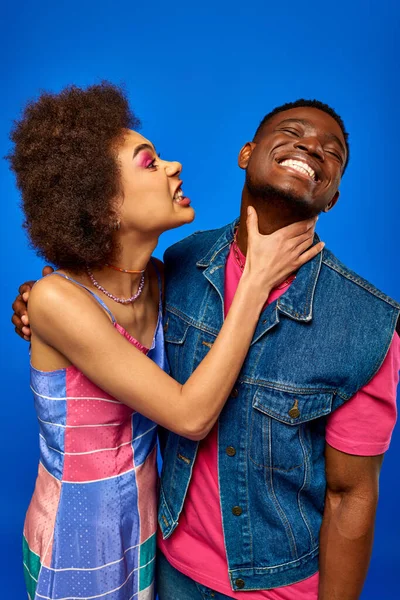 Jeune femme afro-américaine en colère avec un maquillage audacieux touchant le cou du meilleur ami gai dans une tenue d'été élégante tout en se tenant isolé sur bleu, besties à la mode rayonnant de confiance — Photo de stock