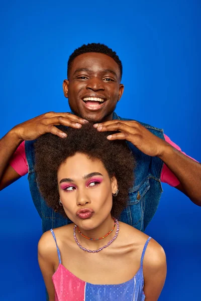 Smiling young african american man with modern hairstyle and summer outfit touching hair of best friend with bold makeup isolated on blue, fashionable besties radiating confidence, friendship — Stock Photo