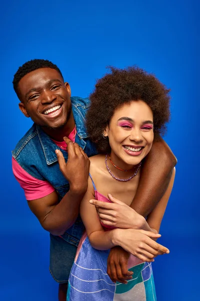 Веселый молодой африканский американец в джинсовом жилете обнимает лучшего друга с смелым макияжем и стильным сарафаном, стоя изолированным на синих, модных подружках, излучающих уверенность — стоковое фото