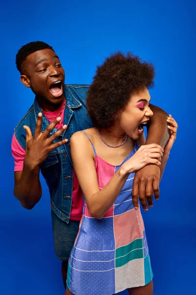 Молодая и модная африканская американка в стильном сарафане кусает руку испуганного лучшего друга в джинсовом жилете и стоит в изоляции на синих, модных подругах, излучающих уверенность, дружбу — стоковое фото