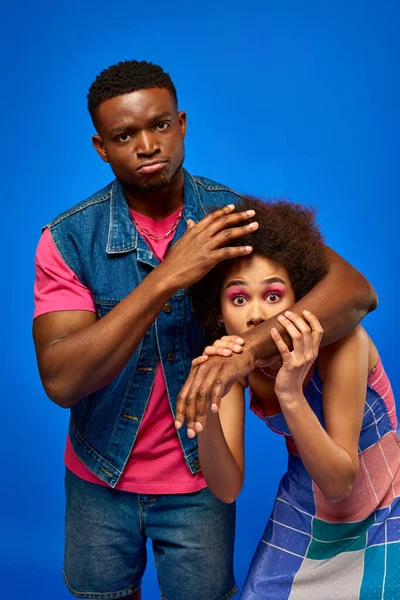 Serious young african american man in summer outfit covering scared best friend in stylish sundress and looking at camera isolated on blue, fashionable besties radiating confidence, friendship — Stock Photo