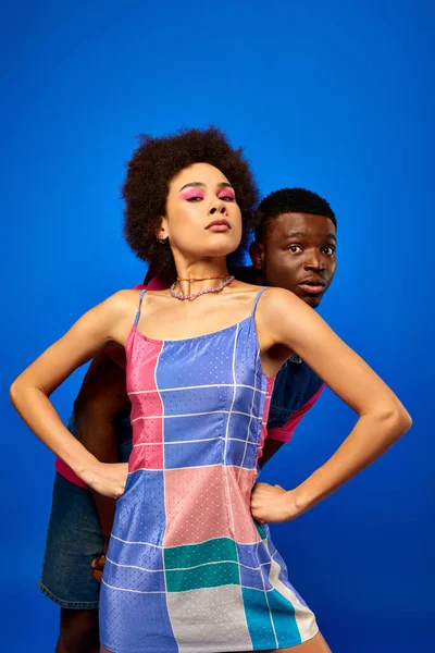 Впевнена молода афроамериканка зі сміливим макіяжем і сарафанчиком тримає руки на стегнах, прикриваючи наляканого найкращого друга ізольованого на синьому, друзі демонструють індивідуальний стиль, дружбу — стокове фото