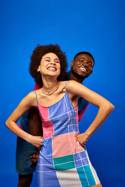 Cheerful young african american woman with bold makeup wearing summer sundress holding hands on hips and looking at camera near best friend isolated on blue, friends showcasing individual style — Stock Photo