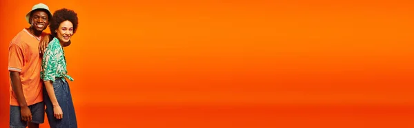 Piacevole giovane uomo afroamericano in panama cappello e vestito estivo guardando la fotocamera mentre in piedi vicino migliore amico con trucco audace isolato su arancione, gli amici in mostra stile individuale, banner — Foto stock