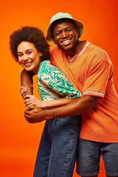 Porträt eines fröhlichen jungen afrikanisch-amerikanischen Mannes mit Panamahut, der seinen besten Freund mit kühnem Make-up umarmt und isoliert auf orange in die Kamera blickt, Freunde, die ihren individuellen Stil präsentieren — Stockfoto
