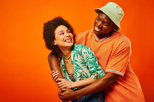 Pleased young african american man in panama hat embracing trendy best friend with bold makeup and looking at each other isolated on orange, friends showcasing individual style — Stock Photo