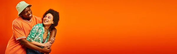 Joven afroamericano alegre en traje de verano y sombrero de panama abrazando mejor amigo con maquillaje audaz y de pie aislado en naranja, amigos mostrando estilo individual, pancarta — Stock Photo