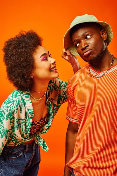 Joven mujer afroamericana alegre con maquillaje audaz tocando el sombrero de panama de la mejor amiga de moda mientras pasa tiempo aislado en naranja, amigos mostrando estilo individual, amistad - foto de stock