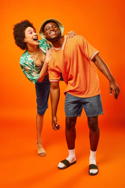 Pleine longueur de jeune femme afro-américaine excitée en tenue d'été étreignant gai meilleur ami dans un chapeau panama tout en se tenant debout sur fond orange, des amis présentant le style individuel — Photo de stock