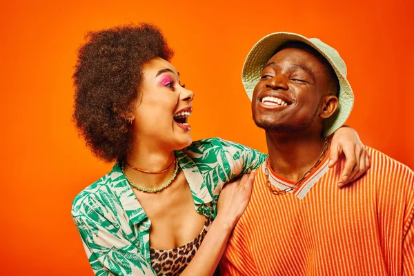 Возбужденная молодая африканская американка с смелым макияжем и модным костюмом, обнимающая лучшего друга в панамской шляпе и стоящая изолированная от оранжевого, друзья демонстрирующие индивидуальный стиль, дружбу — стоковое фото