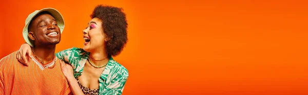 Emocionado joven afroamericana mujer con maquillaje audaz abrazo alegre mejor amigo en traje de verano y sombrero de panama aislado en naranja, amigos mostrando estilo individual, pancarta - foto de stock